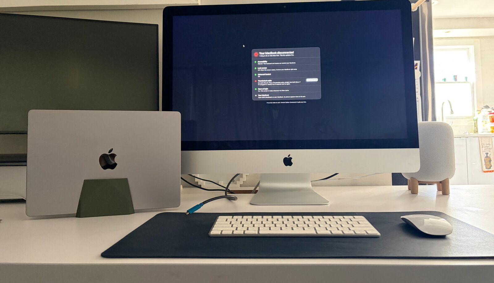 A 27-inch iMac showing the Re5k checklist with green lights for Accessibility, Lock screen, Universal Control and Open at login, and a red light for the Thunderbolt cable, which is lying unplugged in front of it. A closed MacBook stands in a vertical dock beside it.