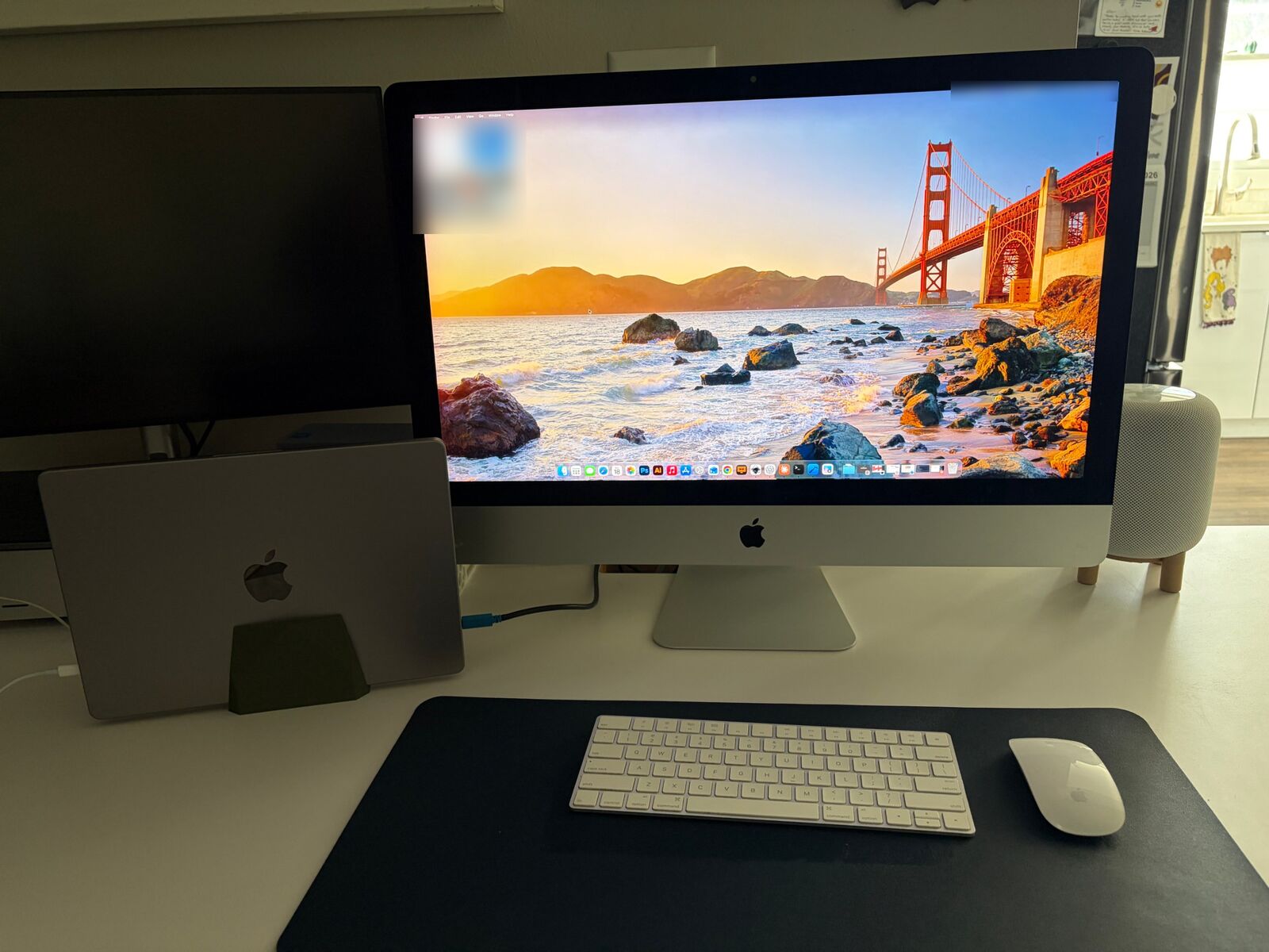 A 2017 27-inch iMac on a desk showing a MacBook's desktop, with the MacBook closed in a vertical stand beside it and the iMac's keyboard and mouse in front.